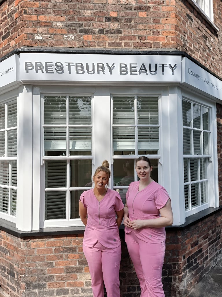 Two beauty salon employees in pink uniforms standing in front of Prestbury Beauty storefront with white trim and brick building