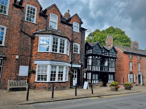 Historic brick and timber-framed buildings on a town street, featuring multi-story residences with bay windows and chimneys under cloudy sky