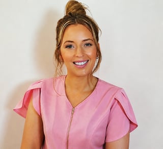Smiling woman wearing a pink zip-up shirt with ruffled sleeves, hair in a top bun, against a white background