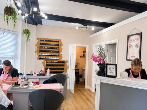 Modern beauty salon interior with two staff members at workstations, wall-mounted product display shelves, and professional styling stations